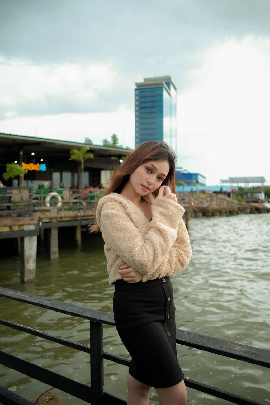A woman in a cozy sweater poses by the river with an urban backdrop. Perfect for lifestyle themes.