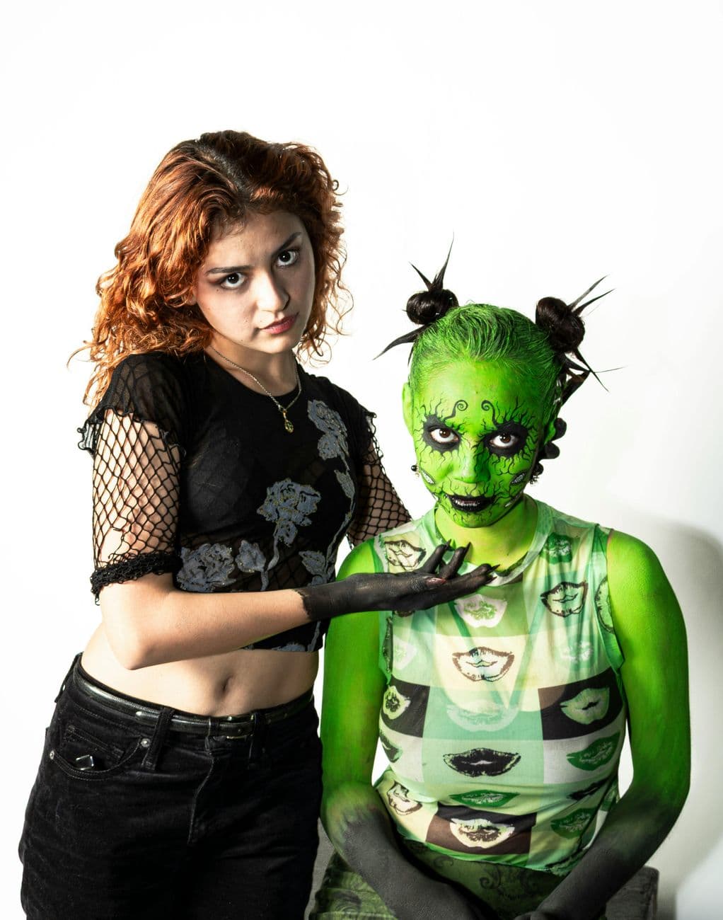 Portrait of two women, one with green artistic makeup and the other with a bold style, against a white background.