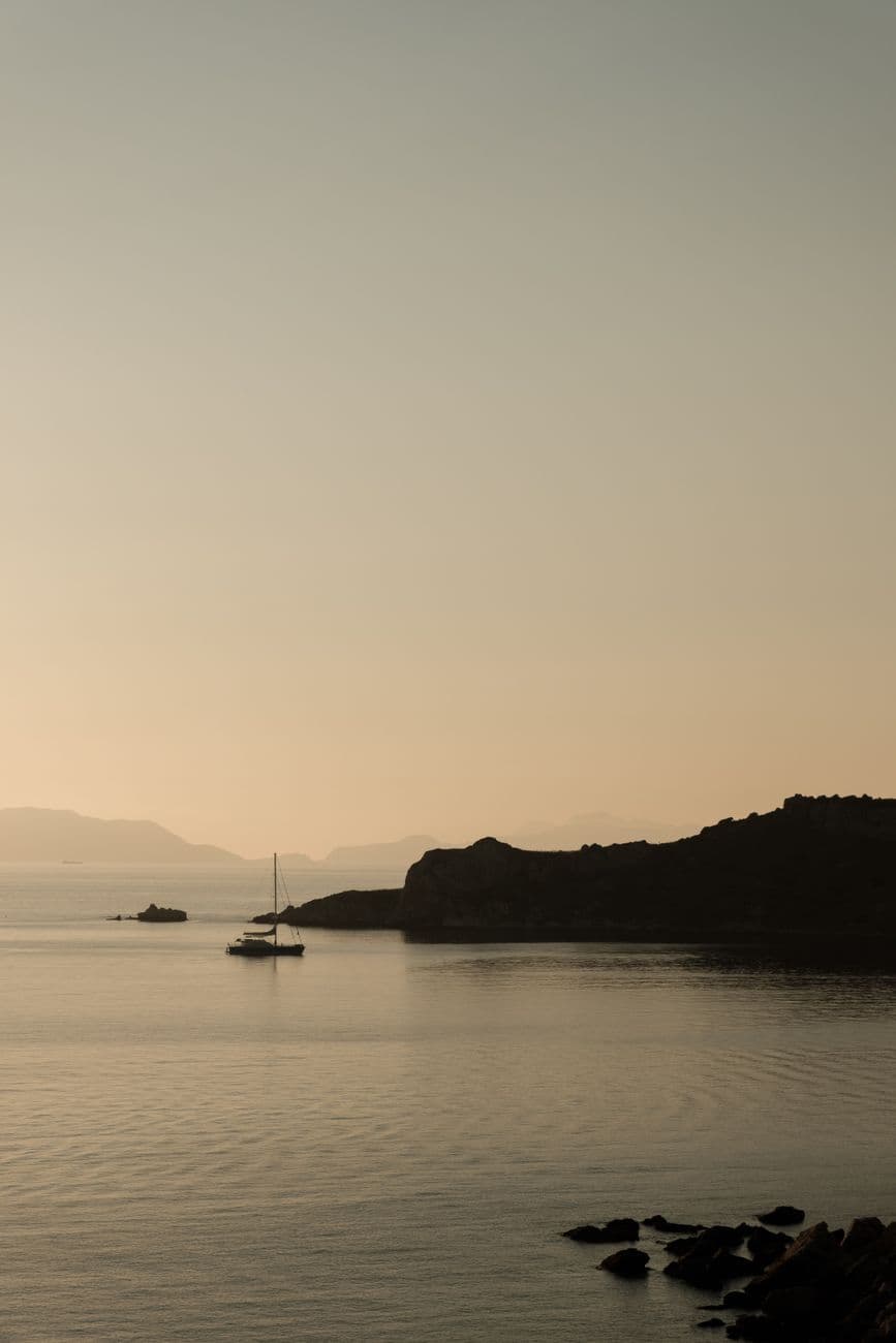 Serene sunset view with sailboat and rocky coastline, casting a tranquil vibe.