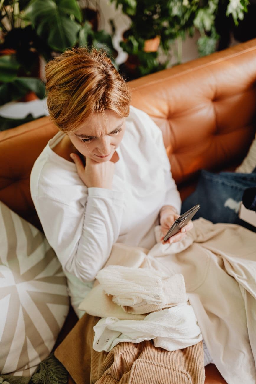 Woman sitting on a sofa looking worried while using her phone, surrounded by clothes indoors.