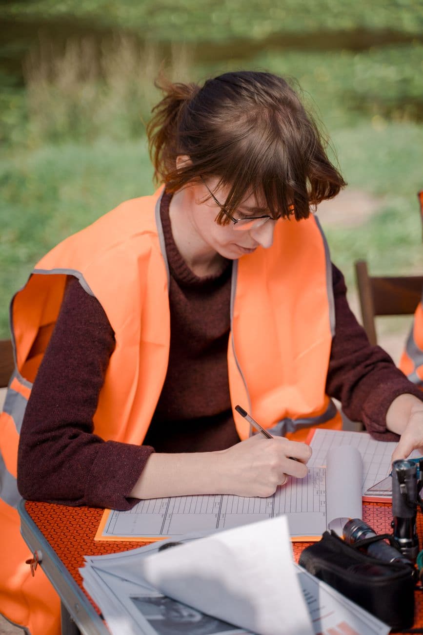 A volunteer in an orange vest documents details at an outdoor event table.