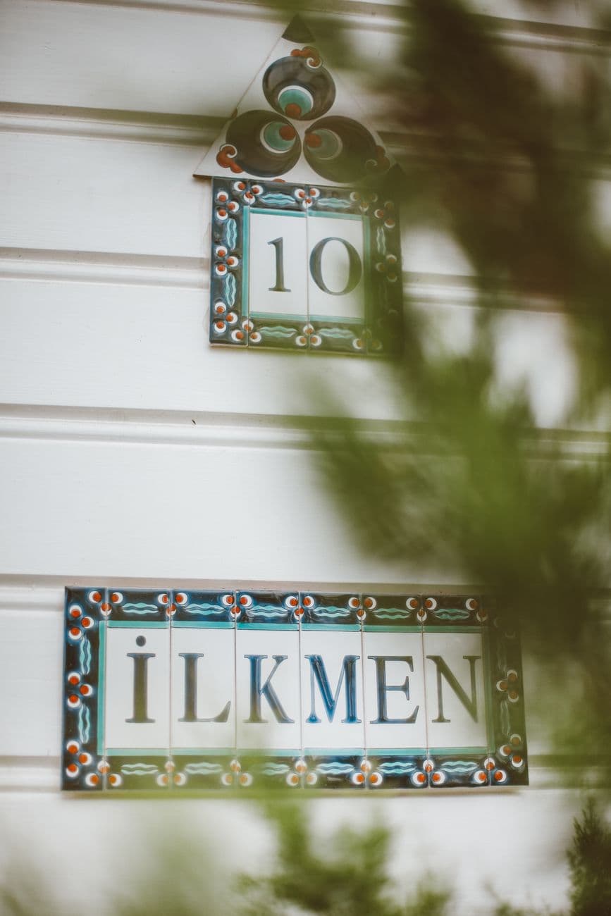 Close-up of decorative house number and name plaques on a white wall, framed by greenery.