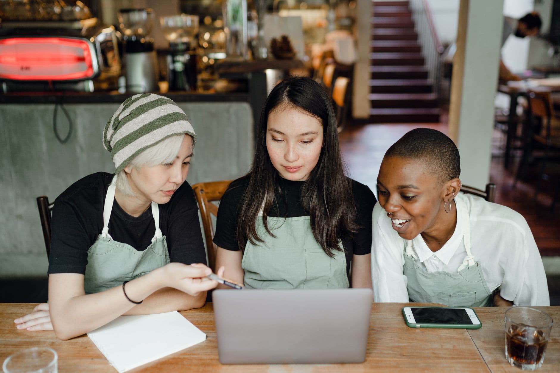 Optimistic smiling diverse female colleagues in aprons browsing netbook and preparing for exam while sitting at table in cozy cafeteria