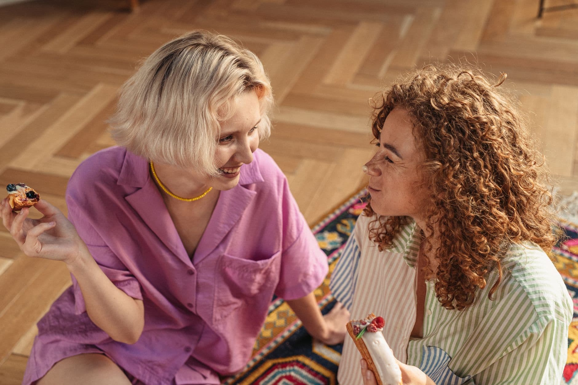 Two women sitting on the floor indoors, sharing desserts and enjoying a warm conversation.