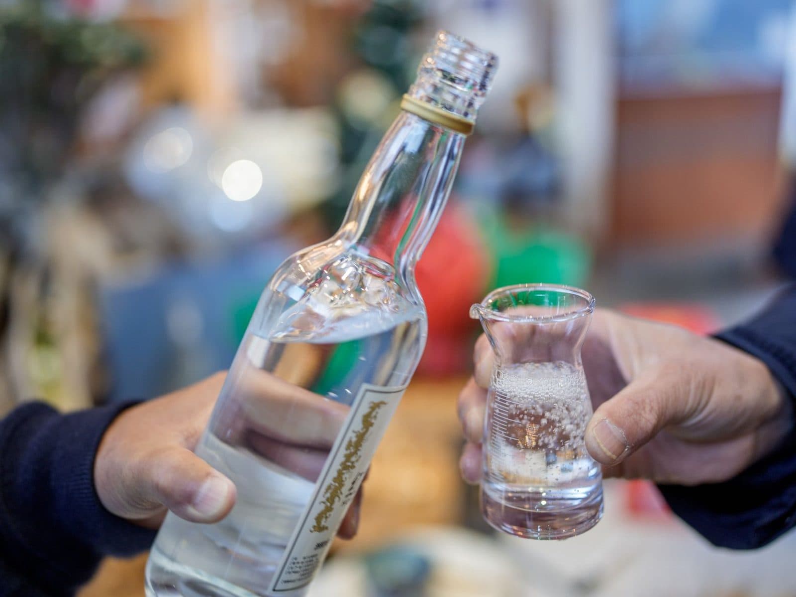 Close-up of hands pouring clear spirits into a shot glass indoors.