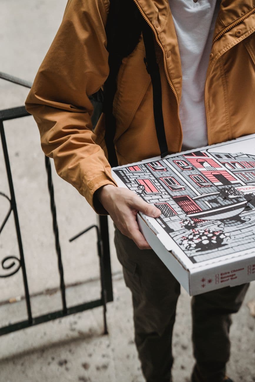Close-up of a delivery person with a pizza box at the doorstep, wearing a brown jacket.