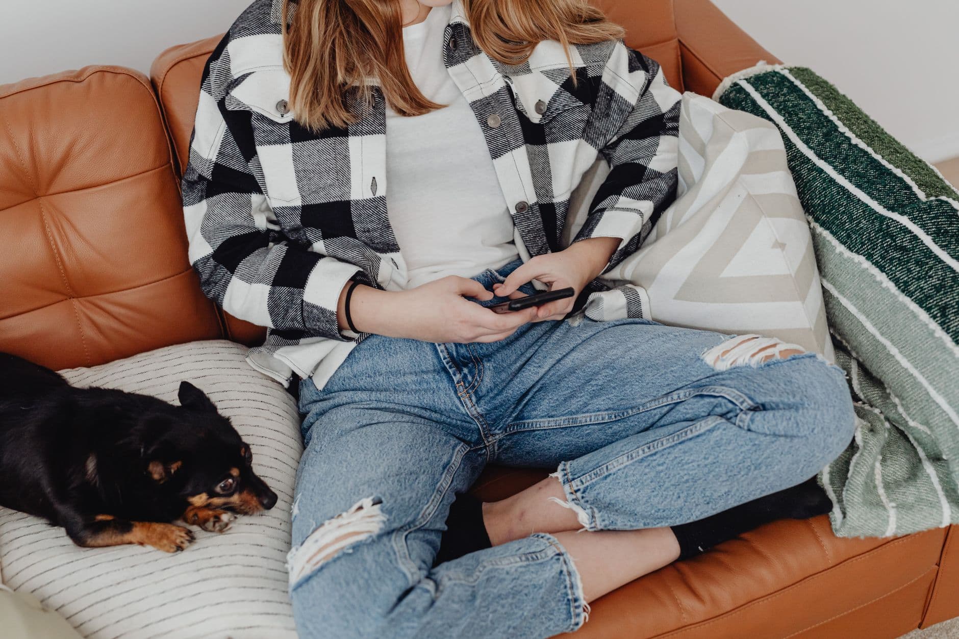 Casual scene of a woman in jeans and plaid shirt using a smartphone with a dog by her side on a cozy sofa.