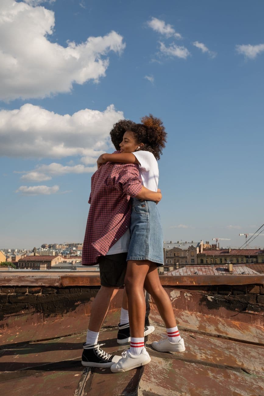 Joyful teenagers hugging on a rooftop with cityscape backdrop under clear blue sky.