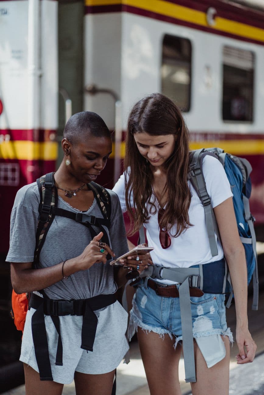 Two female travelers looking at a smartphone while waiting at a train station.