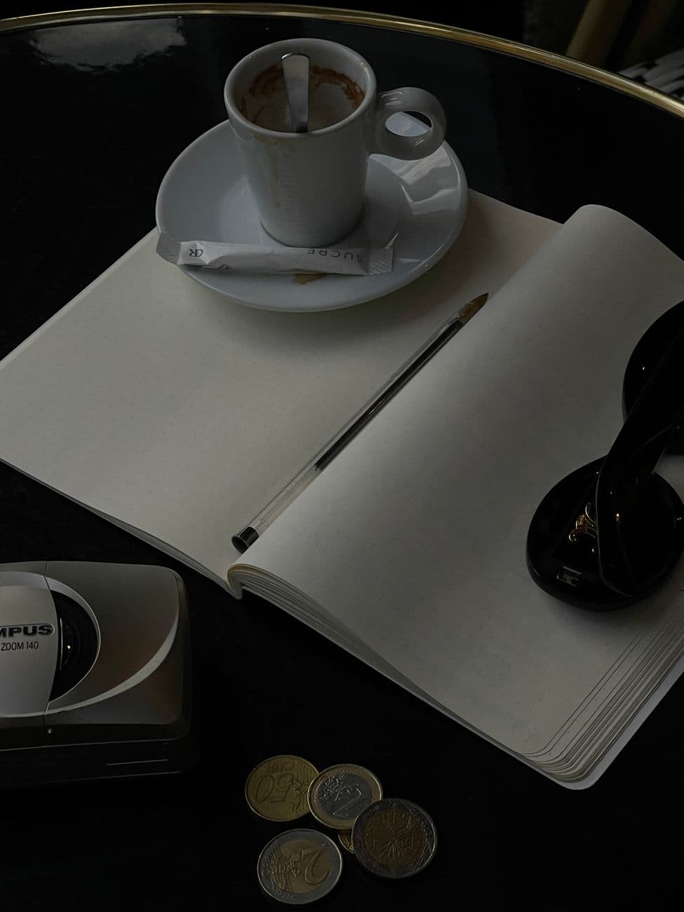 A calming café scene with a coffee cup, open notepad, coins, and sunglasses on a table.
