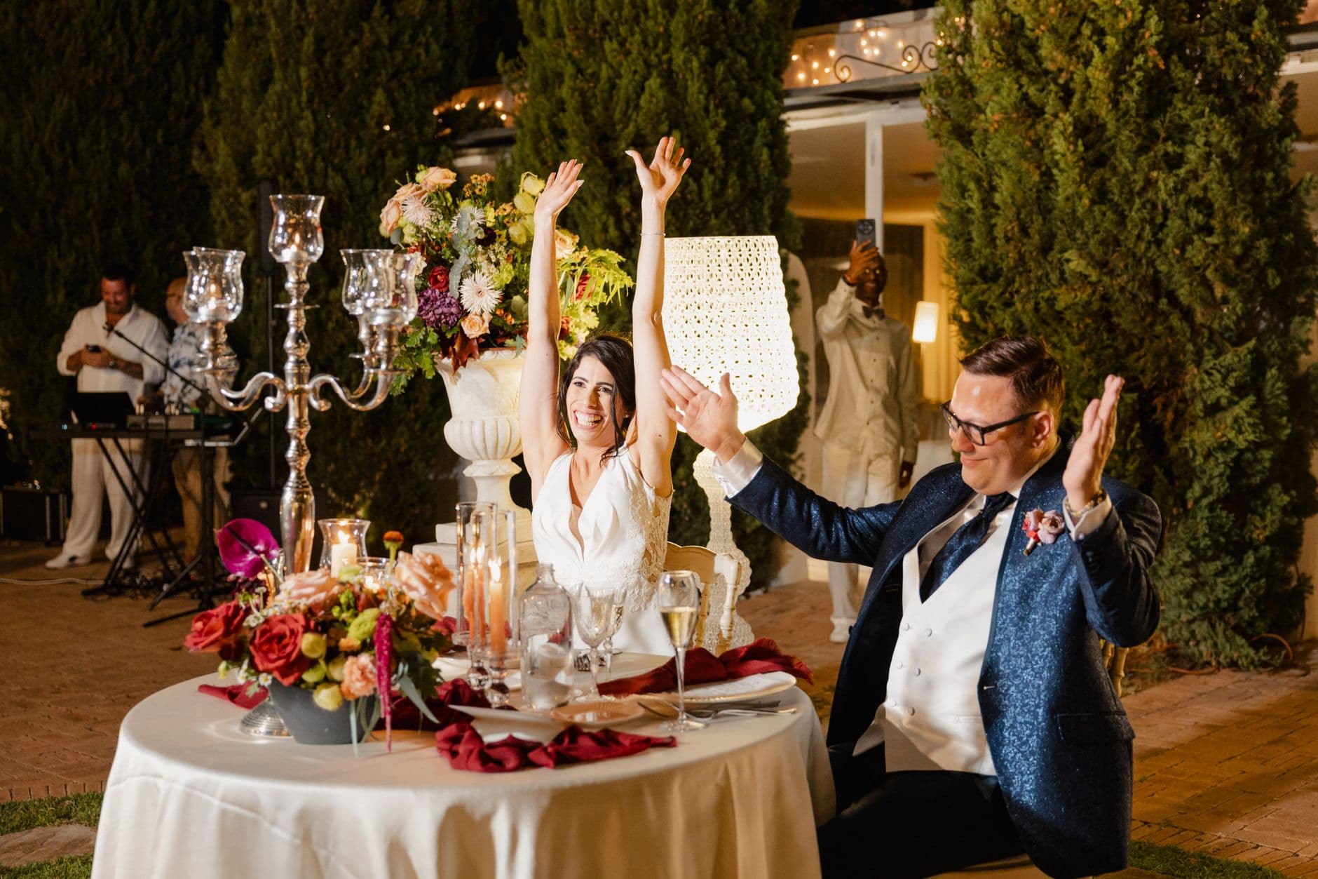 Joyful couple celebrating at an outdoor wedding reception with guests and vibrant floral decorations.