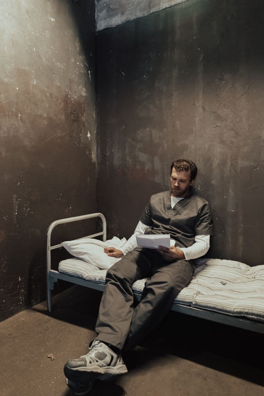 A prisoner in a grungy jail cell reading letters on a small bed, wearing a uniform.