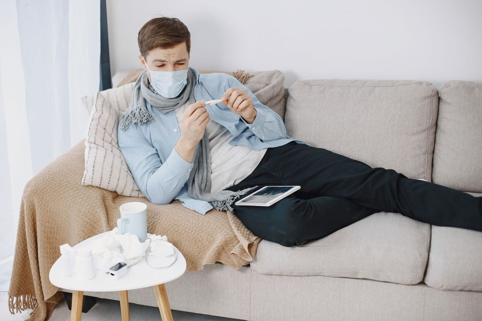 Man wearing a mask checks a thermometer while resting on a couch, surrounded by tissues.