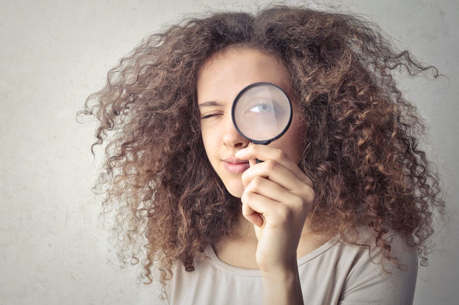 Close-up portrait of a woman winking and holding a magnifying glass against a white background.