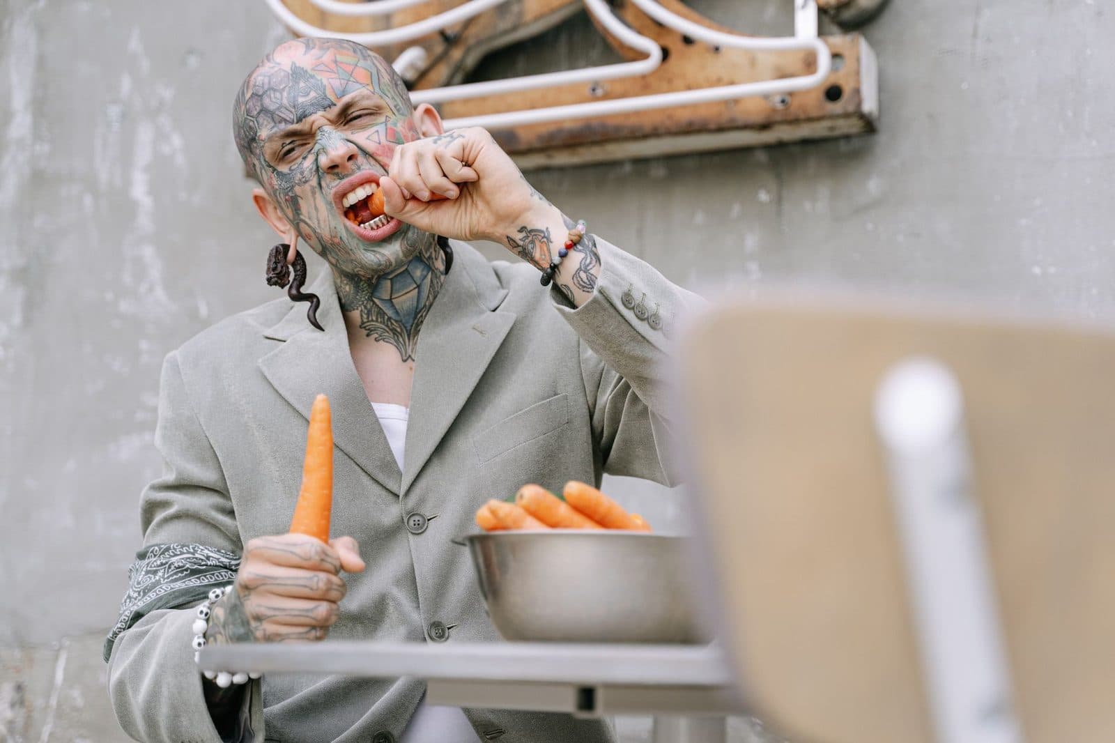 Tattooed man in a gray suit enjoying a carrot outdoors. Perfect for vegan and street style themes.