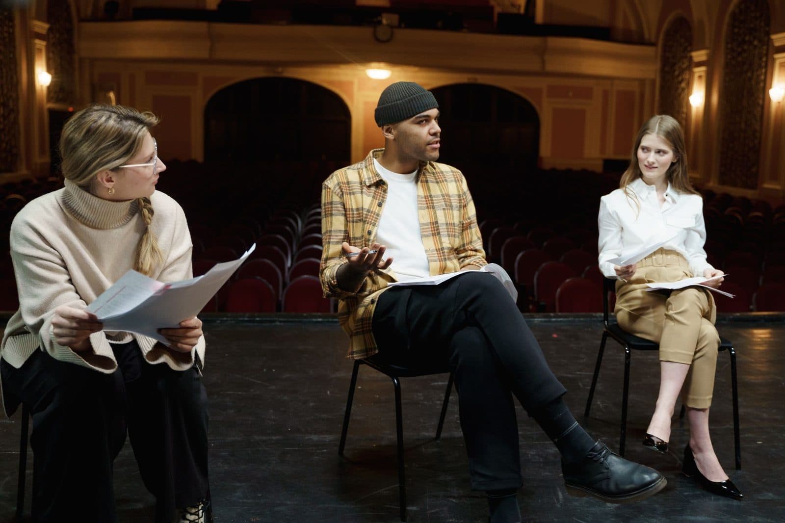 Three actors rehearsing on a theater stage, discussing scripts and performances.