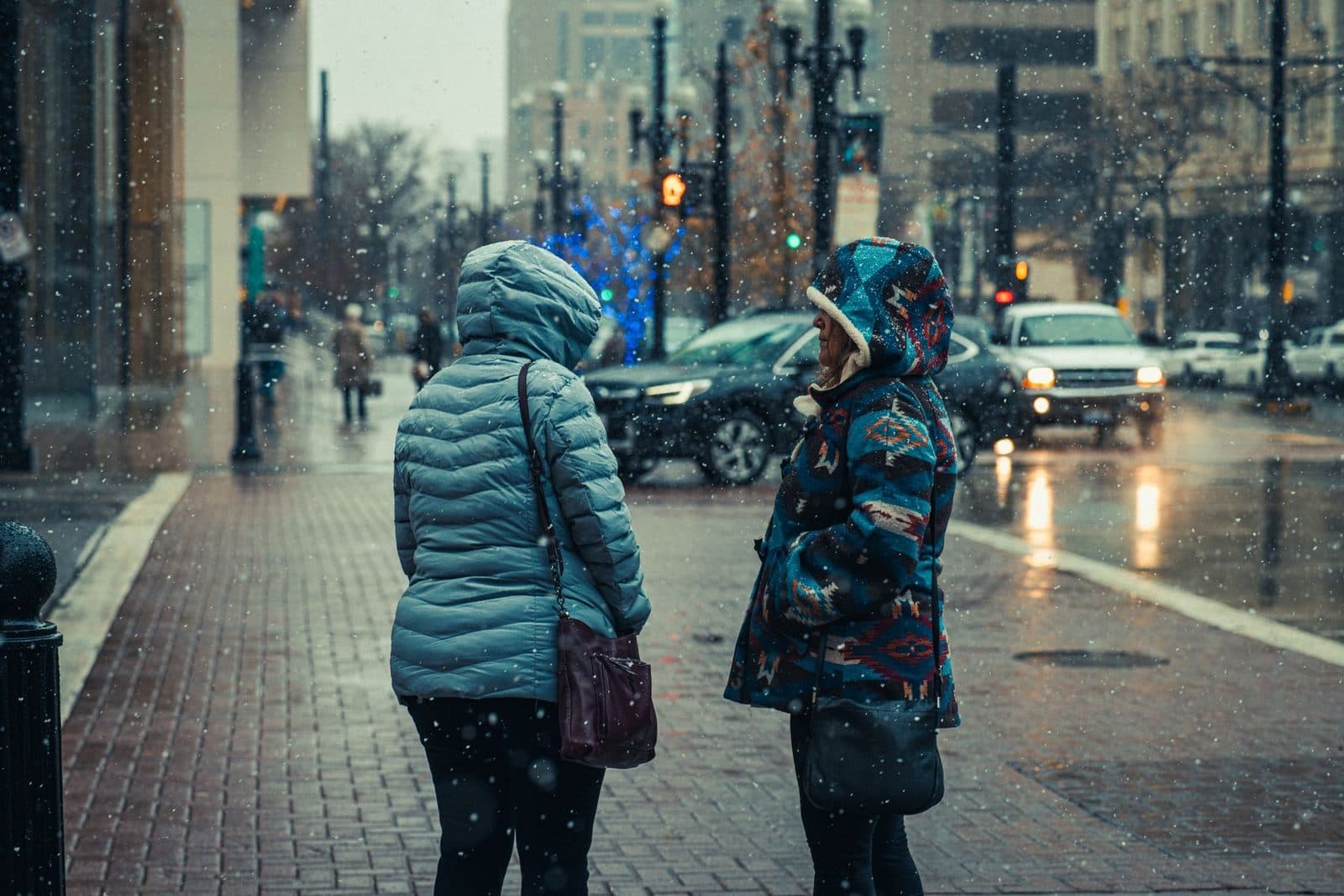 Two women in winter jackets conversing on snow-covered urban sidewalk during the day.