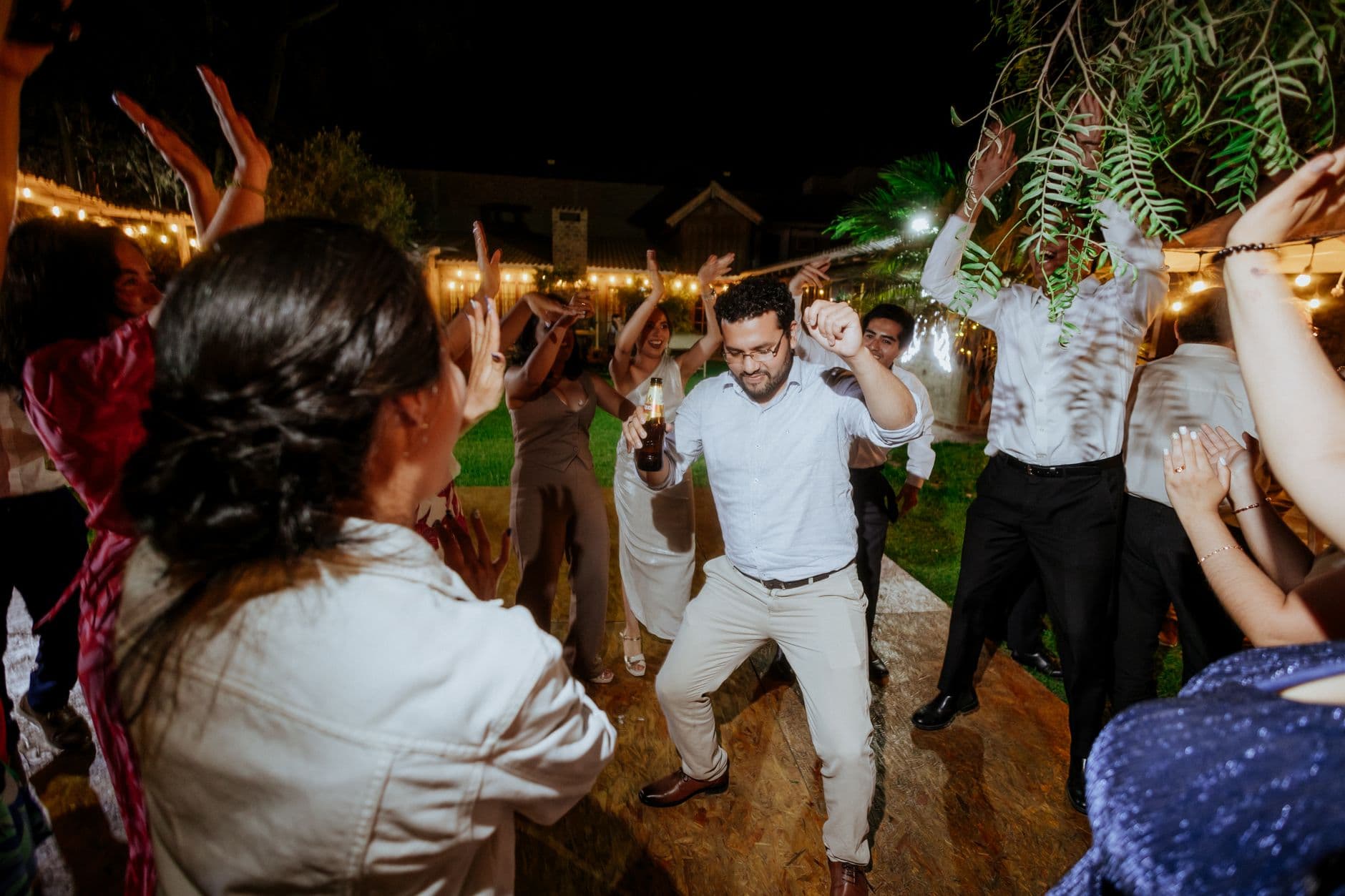 Energetic outdoor wedding reception with people dancing and celebrating under string lights at night.
