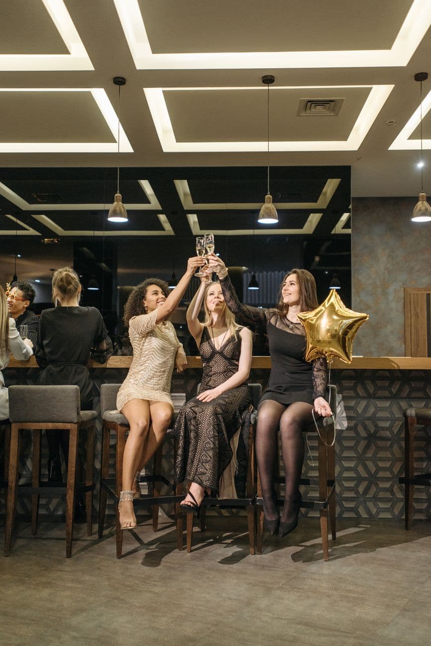 Three women in elegant dresses toasting in a stylish bar setting.