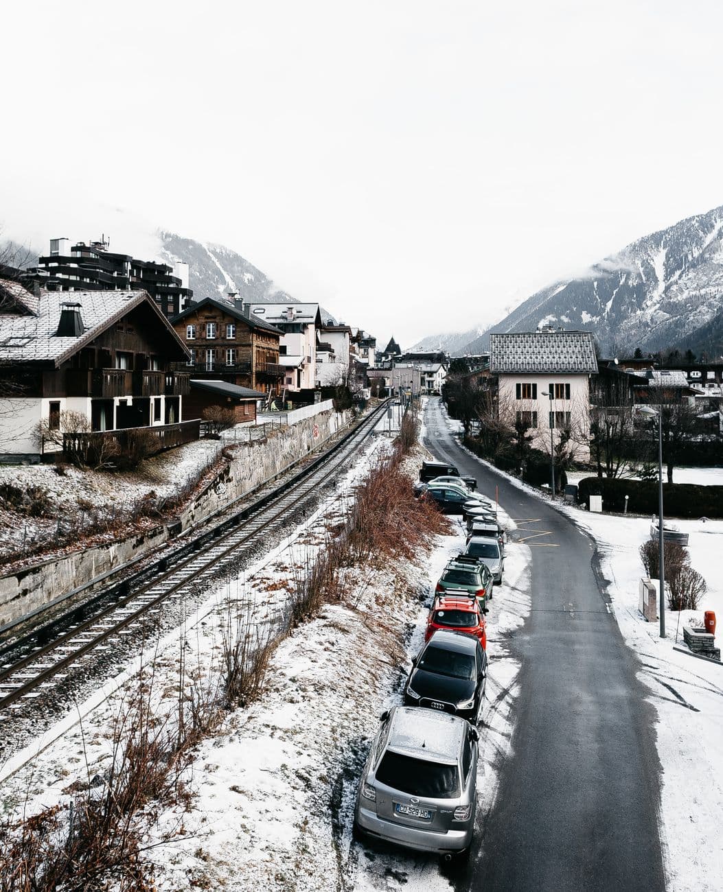 Snow-covered village in Chamonix, France with parked cars along road and railway under winter skies.