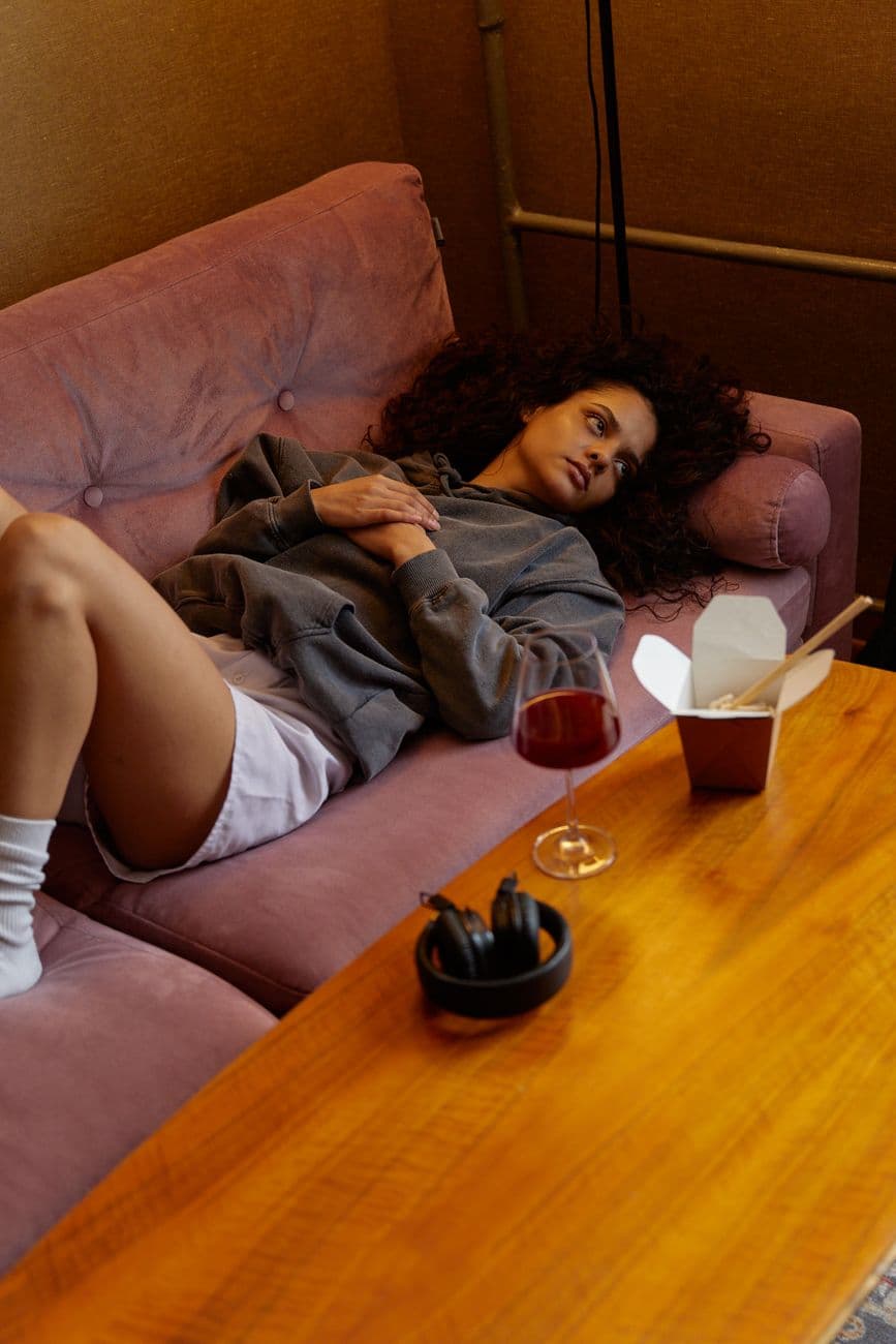 Woman lying on a sofa with a glass of wine and takeout box nearby, depicting relaxation and comfort.