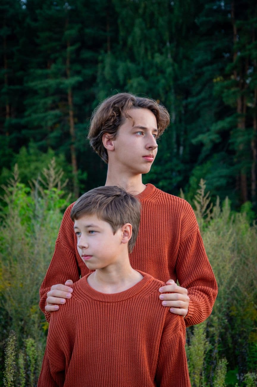 Two brothers in matching sweaters pose outdoors in a lush forest scene.