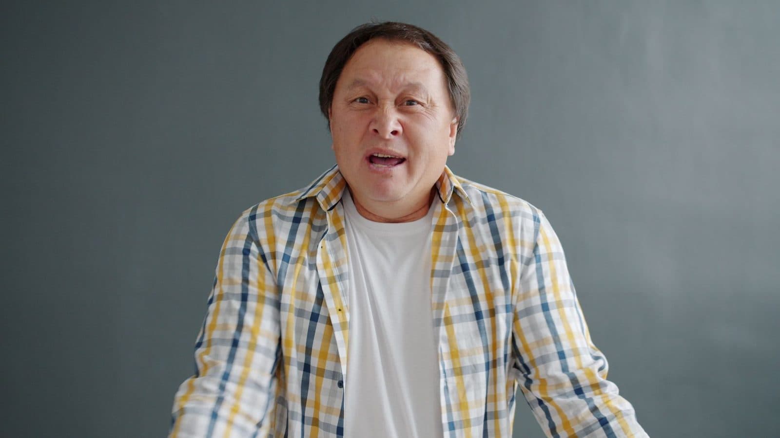 A man in plaid shirt expressing surprise against a gray background.