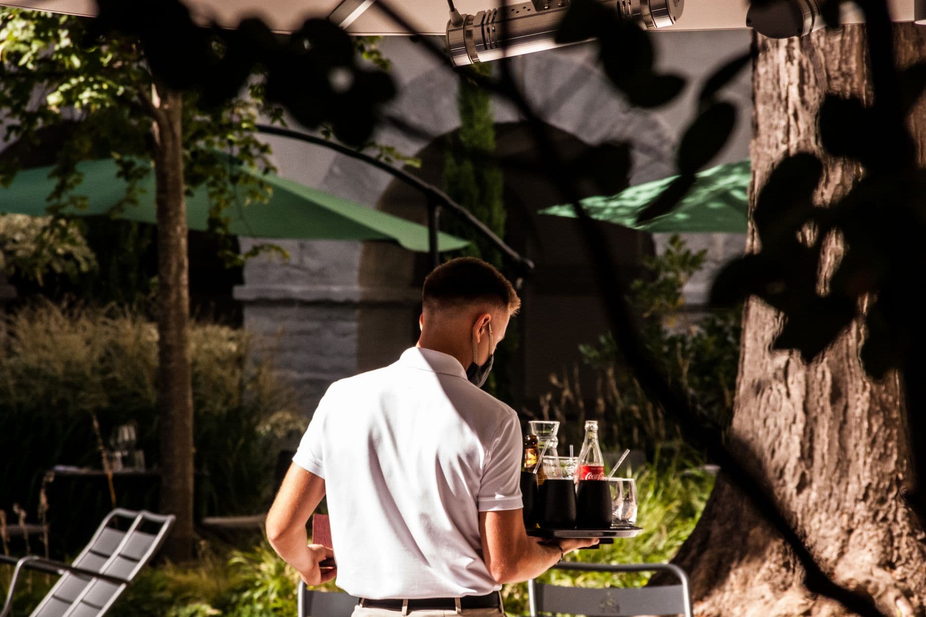 A waiter serves drinks in an outdoor setting, wearing a face mask, embodying the new normal.