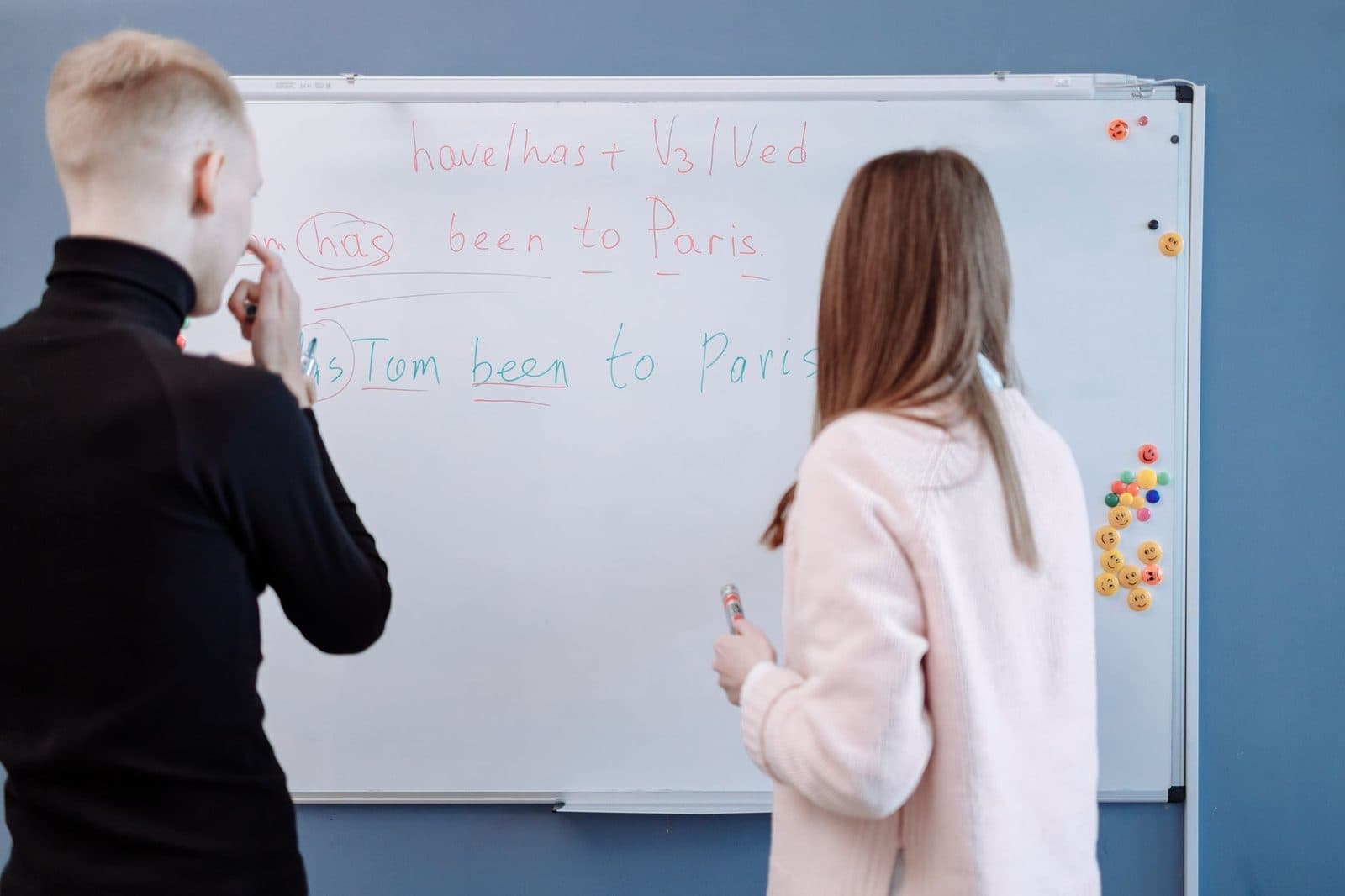 Two students studying English grammar at a whiteboard in a classroom setting.