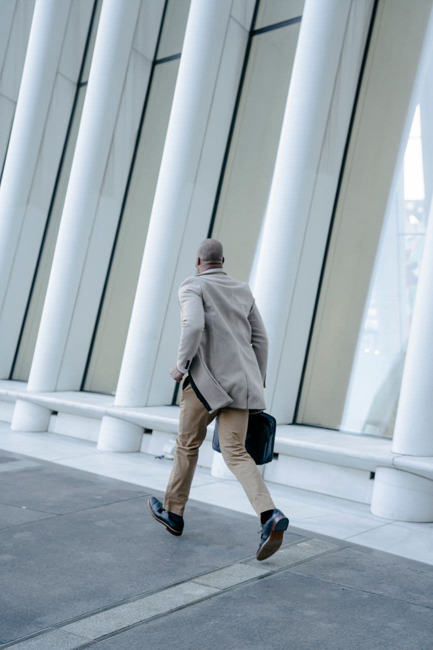 Back view of a man in a brown suit running with a briefcase along architectural columns.